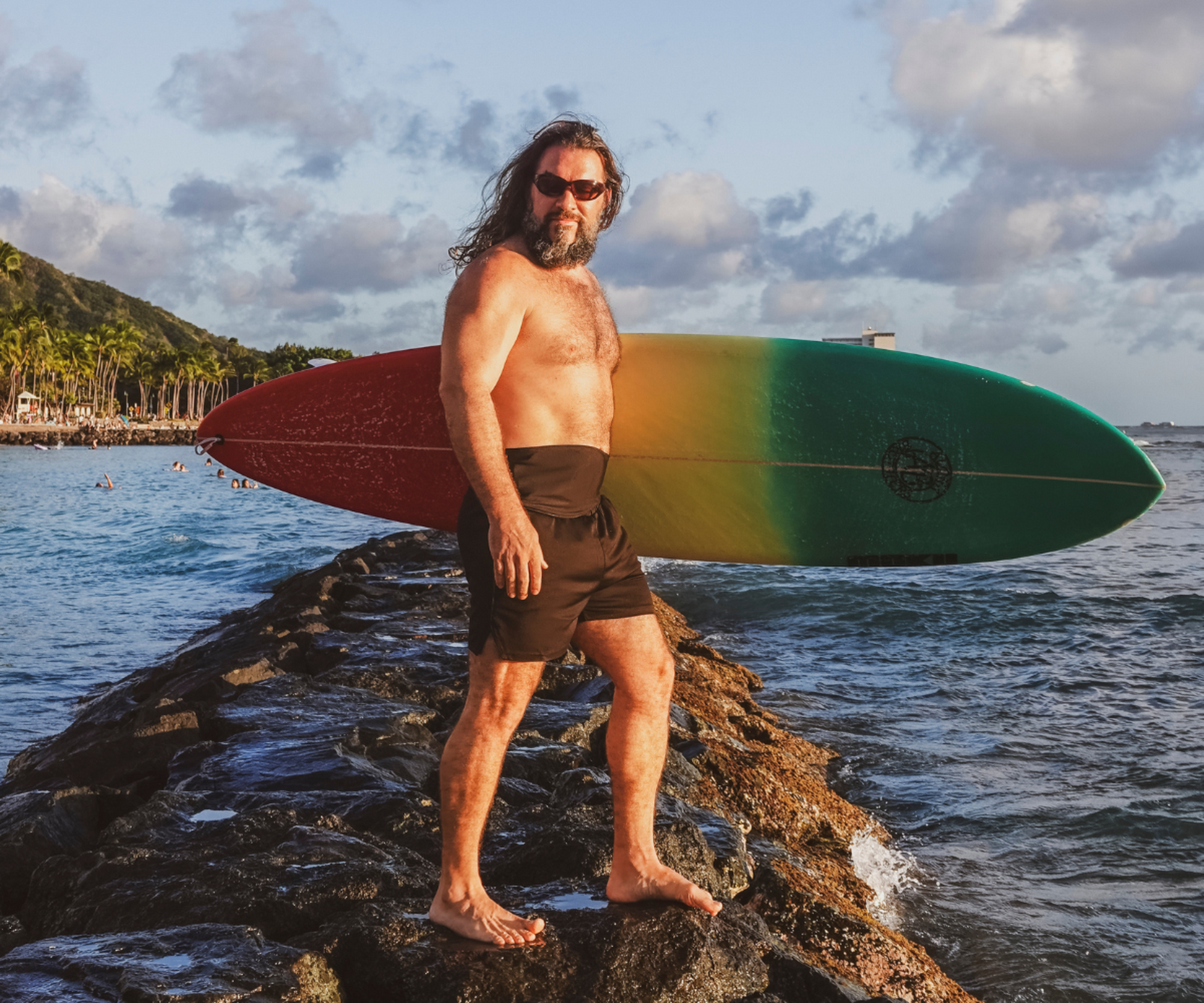 A shirtless man with a beard and sunglasses stands on rocks in the ocean holding a surfboard with a red, yellow, and green gradient. He is wearing dark brown swim trunks, with a tropical shoreline and cloudy sky in the background.